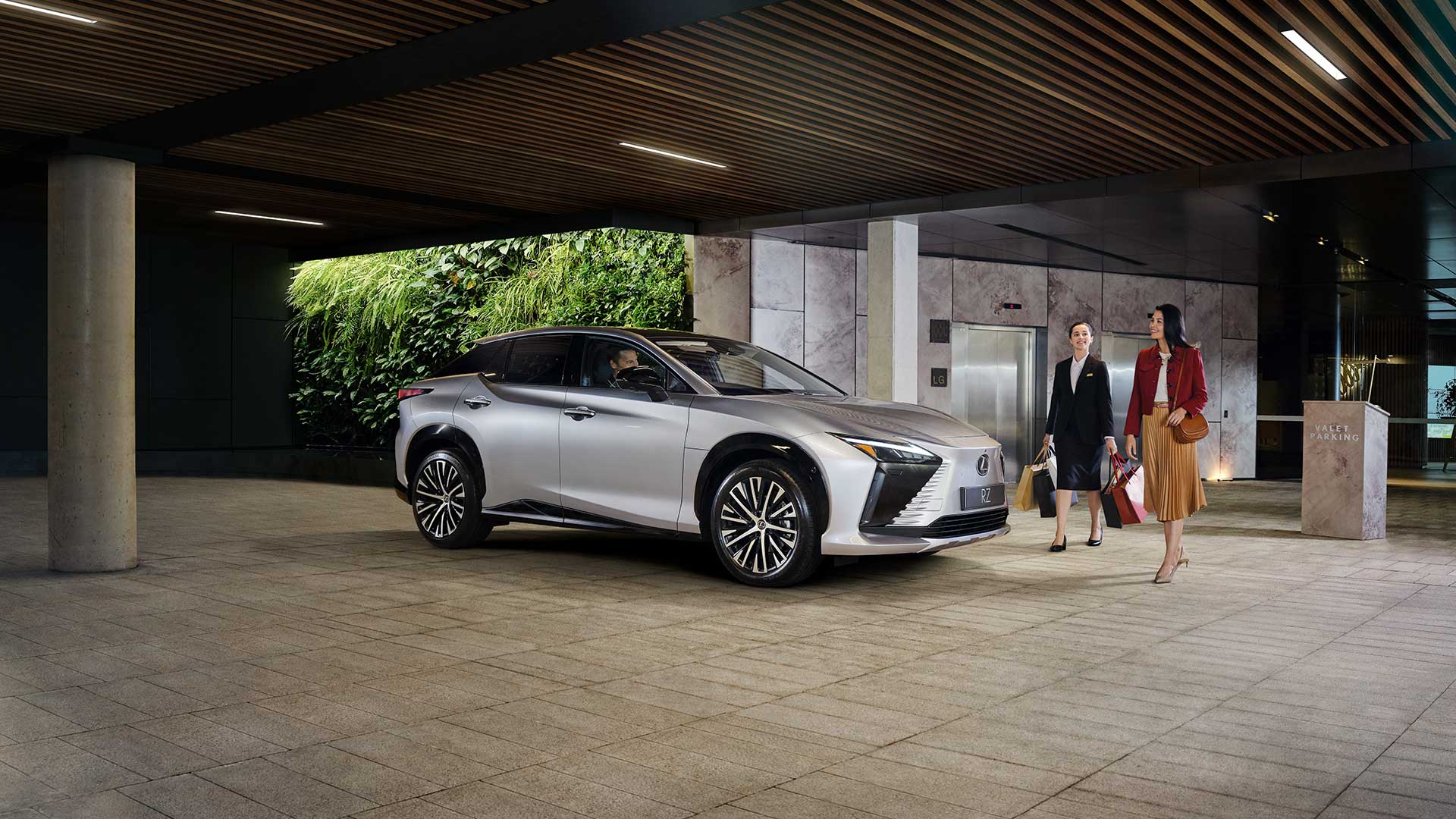 A silver Lexus RZ parked at a valet entrance with two women walking carrying shopping bags.