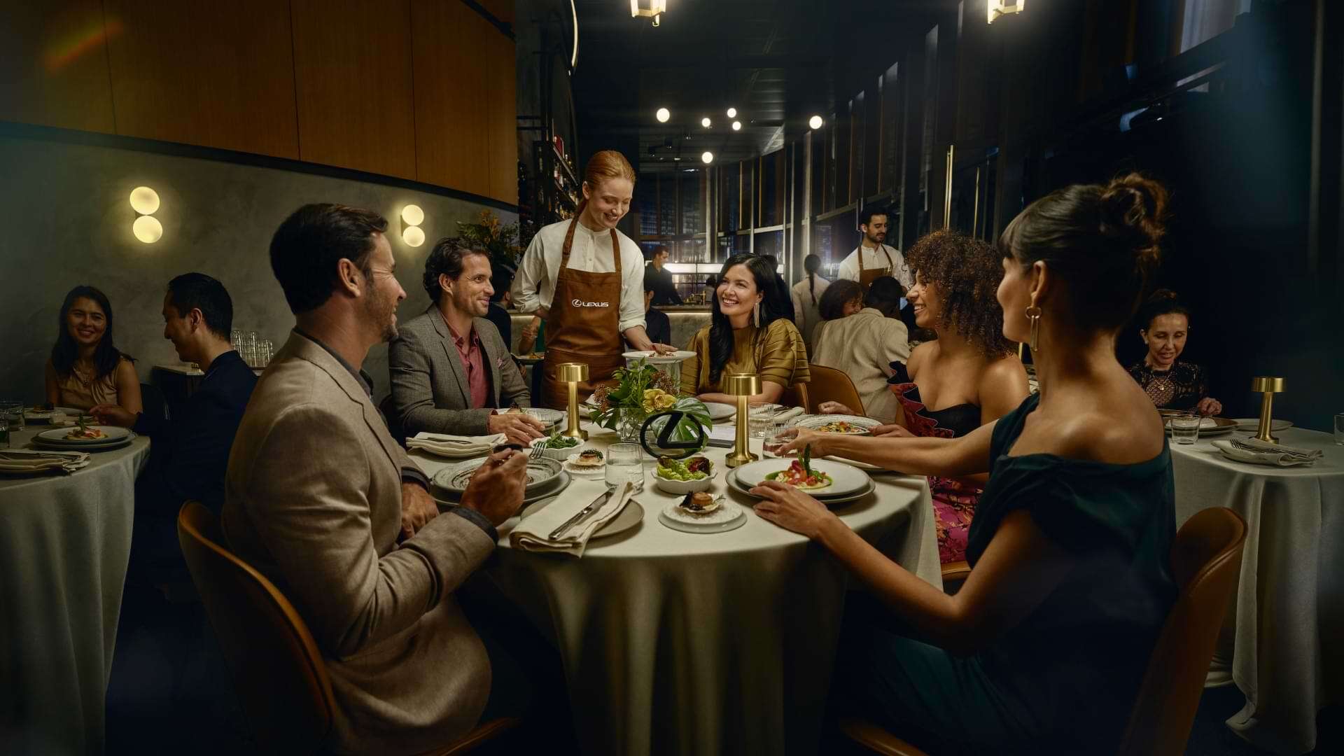 Guests enjoying a fine dining experience as a waiter wearing a Lexus apron serves a dish at a restaurant table.